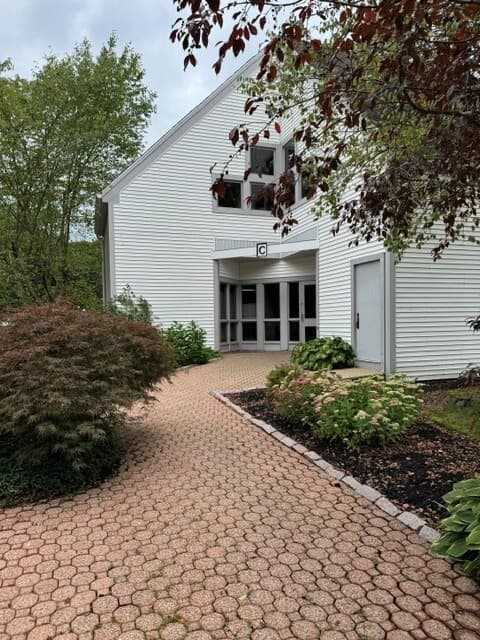 White building entrance beside a brick walkway and landscaped shrubs